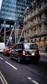 Traffic on a city street with a black taxi leading the line of vehicles. Behind the taxi, there is a red car and a white van. On the left, there's a modern glass building, while on the right an older stone building can be seen, with a Lloyds Bank sign visible.