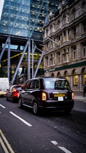 Traffic on a city street with a black taxi leading the line of vehicles. Behind the taxi, there is a red car and a white van. On the left, there's a modern glass building, while on the right an older stone building can be seen, with a Lloyds Bank sign visible.