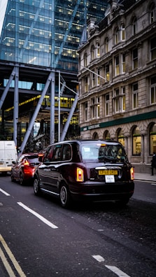 Traffic on a city street with a black taxi leading the line of vehicles. Behind the taxi, there is a red car and a white van. On the left, there's a modern glass building, while on the right an older stone building can be seen, with a Lloyds Bank sign visible.