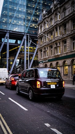 Traffic on a city street with a black taxi leading the line of vehicles. Behind the taxi, there is a red car and a white van. On the left, there's a modern glass building, while on the right an older stone building can be seen, with a Lloyds Bank sign visible.