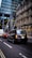 Traffic on a city street with a black taxi leading the line of vehicles. Behind the taxi, there is a red car and a white van. On the left, there's a modern glass building, while on the right an older stone building can be seen, with a Lloyds Bank sign visible.