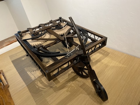 A wooden mechanical device with interlocking gears and pulleys, likely an early machine or model. The structure is set on a wooden surface and appears to be an engineering construct, possibly a historical or replication piece demonstrating mechanics.