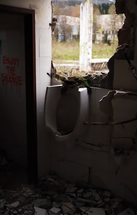 An abandoned bathroom with a urinal partially hidden by debris and a large hole in the wall leading outside. Graffiti on the wall reads 'ENJOY THE SILENCE.' The scene is dimly lit, with broken tiles scattered on the floor, and a view of overgrown vegetation visible through the opening.