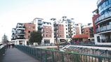 A modern urban residential area featuring multiple mid-rise apartment buildings with balconies and red-brick facades. The setting includes a walkway with a green railing and a few people walking. Street lamps and a small tree are visible, alongside a landscaped area with some plants.