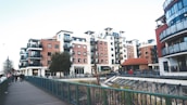 A modern urban residential area featuring multiple mid-rise apartment buildings with balconies and red-brick facades. The setting includes a walkway with a green railing and a few people walking. Street lamps and a small tree are visible, alongside a landscaped area with some plants.