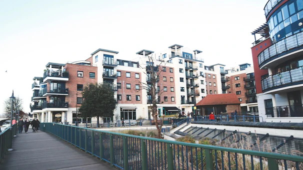 A modern urban residential area featuring multiple mid-rise apartment buildings with balconies and red-brick facades. The setting includes a walkway with a green railing and a few people walking. Street lamps and a small tree are visible, alongside a landscaped area with some plants.