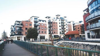 A modern urban residential area featuring multiple mid-rise apartment buildings with balconies and red-brick facades. The setting includes a walkway with a green railing and a few people walking. Street lamps and a small tree are visible, alongside a landscaped area with some plants.