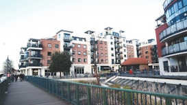 A modern urban residential area featuring multiple mid-rise apartment buildings with balconies and red-brick facades. The setting includes a walkway with a green railing and a few people walking. Street lamps and a small tree are visible, alongside a landscaped area with some plants.