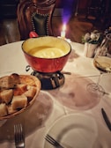 A cozy dining setup featuring a red pot filled with creamy fondue situated on a round table. Nearby, a wicker basket contains sliced bread cubes. A lit candle adds ambiance, and a glass of white wine accompanies the meal. The scene is enhanced by a plush, upholstered chair and subtle decorative elements like a small plant centerpiece.