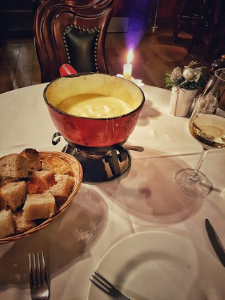 A cozy dining setup featuring a red pot filled with creamy fondue situated on a round table. Nearby, a wicker basket contains sliced bread cubes. A lit candle adds ambiance, and a glass of white wine accompanies the meal. The scene is enhanced by a plush, upholstered chair and subtle decorative elements like a small plant centerpiece.