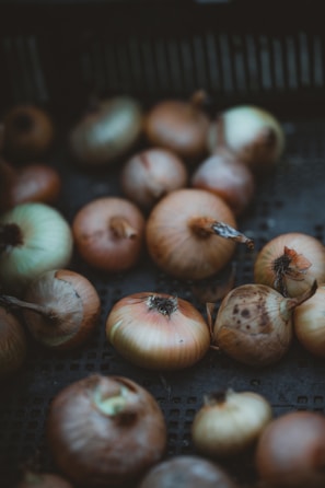 A collection of onions with various shades of brown and yellow rests on a dark surface. The onions have a natural, slightly rustic appearance with some having dried outer skins.
