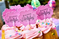 Cupcakes with pink frosting topped with colorful sprinkles and signs that read 'Happy Birthday'. The signs are cute, pink, polka-dotted and flower-shaped. The background includes blurred, colorful, festive decorations.
