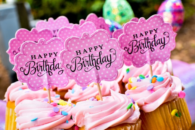 Cupcakes with pink frosting topped with colorful sprinkles and signs that read 'Happy Birthday'. The signs are cute, pink, polka-dotted and flower-shaped. The background includes blurred, colorful, festive decorations.