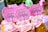 Cupcakes with pink frosting topped with colorful sprinkles and signs that read 'Happy Birthday'. The signs are cute, pink, polka-dotted and flower-shaped. The background includes blurred, colorful, festive decorations.