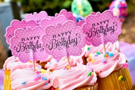 Cupcakes with pink frosting topped with colorful sprinkles and signs that read 'Happy Birthday'. The signs are cute, pink, polka-dotted and flower-shaped. The background includes blurred, colorful, festive decorations.
