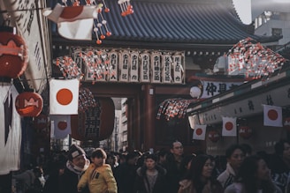 A bustling street scene in Japan with a large crowd of people walking beneath traditional architecture adorned with Japanese flags. Lanterns and decorated strings with red and white colors hang from the rooftops, creating a festive atmosphere. The setting appears to be a market or festival environment, with numerous individuals interacting in a lively mood.