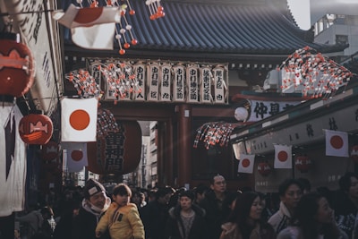 A bustling street scene in Japan with a large crowd of people walking beneath traditional architecture adorned with Japanese flags. Lanterns and decorated strings with red and white colors hang from the rooftops, creating a festive atmosphere. The setting appears to be a market or festival environment, with numerous individuals interacting in a lively mood.