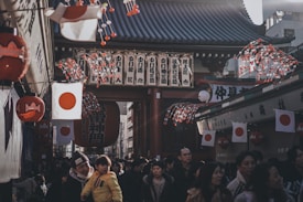 A bustling street scene in Japan with a large crowd of people walking beneath traditional architecture adorned with Japanese flags. Lanterns and decorated strings with red and white colors hang from the rooftops, creating a festive atmosphere. The setting appears to be a market or festival environment, with numerous individuals interacting in a lively mood.