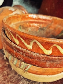 A close-up view of stacked earthenware bowls with intricate, painted designs. The bowls display a rustic finish and feature wavy yellow patterns and earthy tones. The surface appears textured and well-used, suggesting they might be handmade pottery.