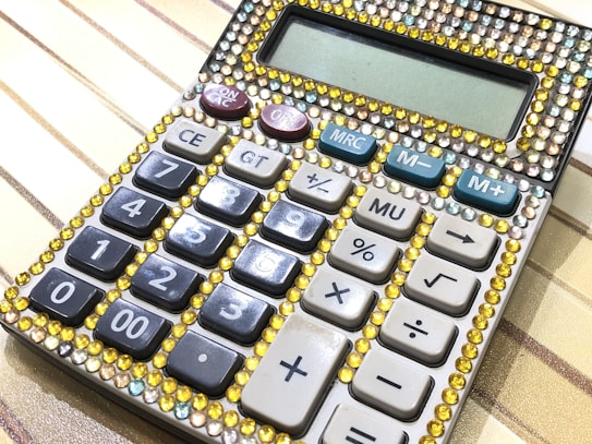 A close-up view of a calculator embellished with colorful rhinestones. The rhinestones are predominantly yellow with some light blue, creating a decorative frame around the display and buttons. The calculator features standard numeric keys, function keys, and special buttons such as 'M+', 'M-', 'MU', and 'GT'. The background surface has a striped pattern with shades of beige and gold.