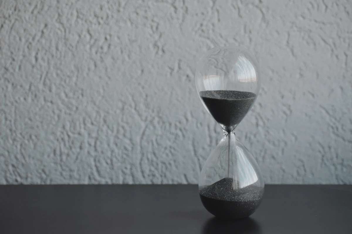 A glass hourglass with dark sand on a black table against a textured grey wall