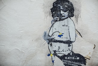 Street art of a child figure holding a flower on a textured wall. The artwork, predominantly in black and white, features accents of blue and yellow. Text reading 'System Change Not Climate Change' is visible near the bottom.