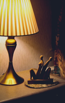 A warmly lit lamp sits on a table beside a stylized wooden sculpture depicting abstract human figures. The light from the lamp casts a soft glow on the textured wall in the background, creating a cozy and inviting ambiance.