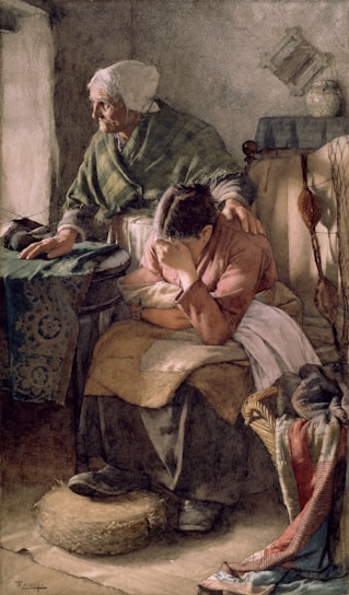 An elderly woman with a shawl comforts a younger woman who is seated and appears distressed, covering her face with her arm. The scene is set in a modest, dimly lit room with simple furnishings and textiles.