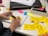 A person is writing on a yellow sticky note with a black pen. Several sticky notes with handwritten text in French are spread on a white table. The person is sitting beside a laptop, and there are colorful sticky notes in the background along with a notebook and a pen.