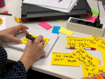 A person is writing on a yellow sticky note with a black pen. Several sticky notes with handwritten text in French are spread on a white table. The person is sitting beside a laptop, and there are colorful sticky notes in the background along with a notebook and a pen.