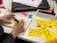 A person is writing on a yellow sticky note with a black pen. Several sticky notes with handwritten text in French are spread on a white table. The person is sitting beside a laptop, and there are colorful sticky notes in the background along with a notebook and a pen.