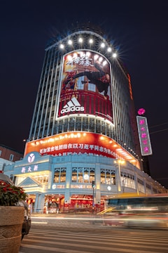 A bustling cityscape featuring a tall, illuminated building with a large Adidas advertisement. The scene is enhanced by bright lights and the motion blur of passing vehicles on the street below. The ground floor includes a Burger King, and the surrounding area is lively with vibrant signage in Chinese characters.