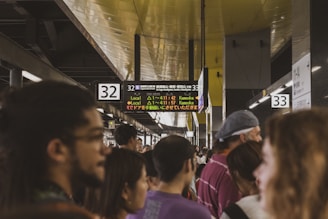 A crowded train station platform with people waiting under illuminated signs indicating train schedules and platforms. The ceiling is lined with lights and the signs display information in both English and Japanese.
