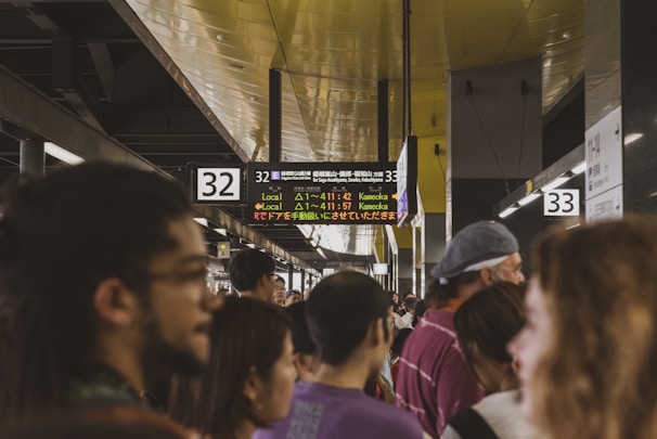 A crowded train station platform with people waiting under illuminated signs indicating train schedules and platforms. The ceiling is lined with lights and the signs display information in both English and Japanese.