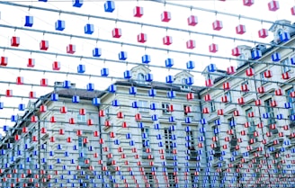 An installation of numerous hanging rectangular objects in red and blue colors is suspended in the foreground, contrasting against a classic European-style building with ornate windows and a tiled roof in the background.