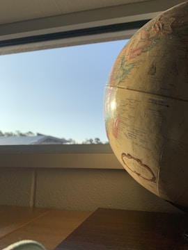A globe is positioned near a window with sunlight streaming in, casting a warm glow over its surface. The sky outside is clear with a subtle blue hue. The interior appears to be a room with neutral colored walls and a wooden surface beneath the globe.