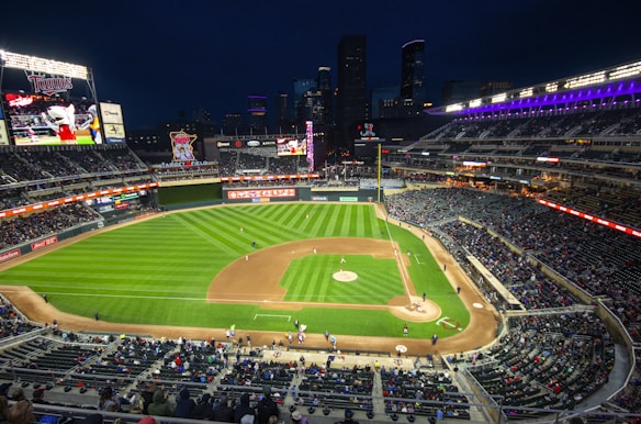 A baseball game is taking place in a large, brightly lit stadium during nighttime. The field is well-maintained with a distinct diamond shape and lush green grass. The stands are filled with spectators, and large screens display graphics and video footage related to the game. The city skyline is visible in the background.