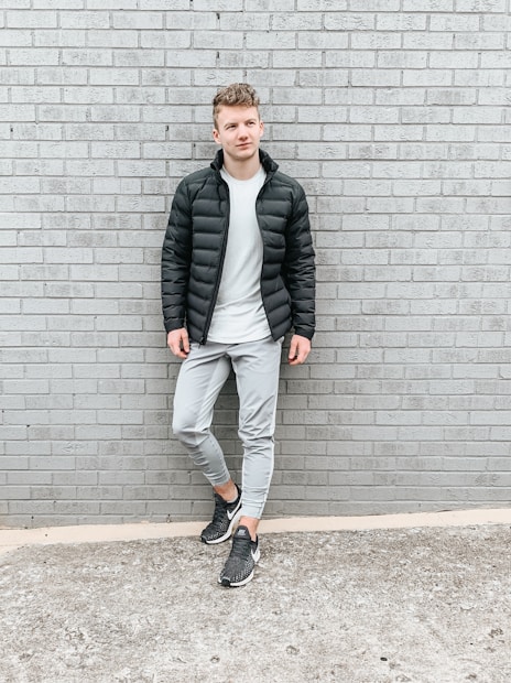 A person stands casually against a gray brick wall wearing a black puffer jacket, a white t-shirt, light gray pants, and black sneakers. The setting appears urban with a minimalist style.