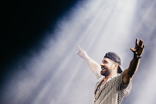 A man with a beard, wearing a striped shirt and a backwards cap, stands with his arms outstretched, smiling broadly under a spotlight. The background is dark and smoky, highlighting the subject under the bright stage light.