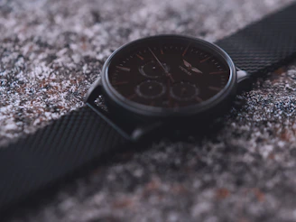 Close-up of a sleek men's watch with a dark metal strap resting on a textured black surface.