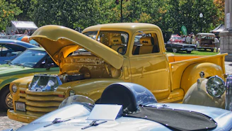 A close-up view of a vibrant yellow vintage truck with its hood open, showcasing the engine. The interior seats feature a batman logo. Surrounding the yellow truck are other classic cars in various colors, parked in an outdoor area with trees and people in the background.