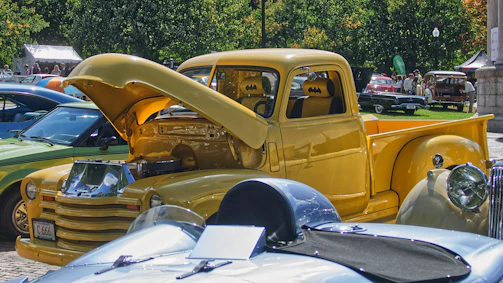 A close-up view of a vibrant yellow vintage truck with its hood open, showcasing the engine. The interior seats feature a batman logo. Surrounding the yellow truck are other classic cars in various colors, parked in an outdoor area with trees and people in the background.