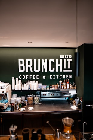 A modern coffee bar setup with a range of coffee-making equipment and supplies. The background displays a prominent sign with 'BRUNCHIT Coffee & Kitchen' in bold white letters. Various coffee cups, jars, bottles, and a professional espresso machine are arranged neatly on the counter.