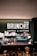 A modern coffee bar setup with a range of coffee-making equipment and supplies. The background displays a prominent sign with 'BRUNCHIT Coffee & Kitchen' in bold white letters. Various coffee cups, jars, bottles, and a professional espresso machine are arranged neatly on the counter.
