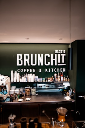A modern coffee bar setup with a range of coffee-making equipment and supplies. The background displays a prominent sign with 'BRUNCHIT Coffee & Kitchen' in bold white letters. Various coffee cups, jars, bottles, and a professional espresso machine are arranged neatly on the counter.