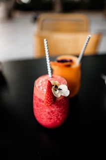 Two colorful drinks are placed on a dark surface. The drink in the foreground is a vibrant pink, topped with a strawberry and a white flower, and has a striped straw. The background features an orange drink with a similar straw, possibly garnished with edible flowers.