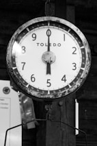 A vintage hanging scale with a round face displaying numbers 1 through 9 and marked with increments for precision. The brand 'Toledo' is prominently mentioned in the center. The scale has an aged appearance with some rust visible on the metallic parts, set against a wooden background.