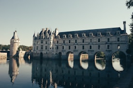 A historic château with an elegant Renaissance architectural style is seen spanning a river. The building's arches allow water to flow underneath, and its reflection is visible on the water's surface. Adjacent towers and a pathway lined with trees enhance the picturesque scene.