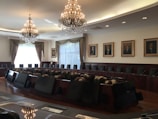 A large, elegant conference room featuring a long, oval table surrounded by high-backed chairs. Two impressive chandeliers hang from the ceiling, casting a warm glow. The walls are adorned with framed portraits, and large windows with curtains allow natural light to enter the room.