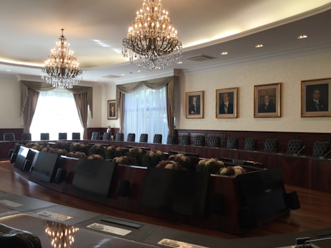 A large, elegant conference room featuring a long, oval table surrounded by high-backed chairs. Two impressive chandeliers hang from the ceiling, casting a warm glow. The walls are adorned with framed portraits, and large windows with curtains allow natural light to enter the room.